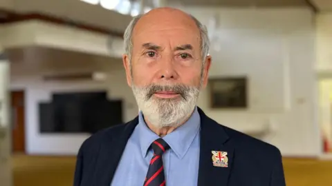 A man with a grey beard in a blue suit and a stripped tie.