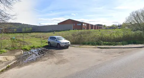 A field is separated from the road by a wire fence. Trees and sparse hedges are dotted along the edge. A long brick building is on the other side of the field. A car is parked away from the road, next to the field.