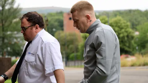 Cameron Jones being led into court, wearing a grey jacket and handcuffed to a guard in a white shirt and black tie