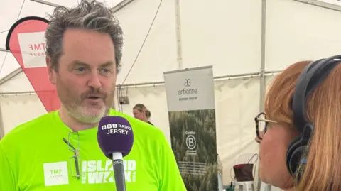 A woman wearing headphones and a striped shirt holds a microphone up to a man in a neon yellow 'officials' T-shirt as the pair chat about the race