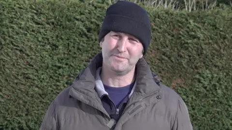 A man wearing a black woolly hat and grey coat, stood in front of a green bush and looking to the camera.