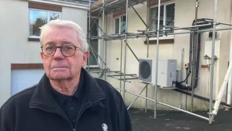 A man with glasses is looking straight at the camera - he has short white hair and is wearing a black fleece.  Behind him is house with scaffolding surronding a heat pump which is mounted on the wall.