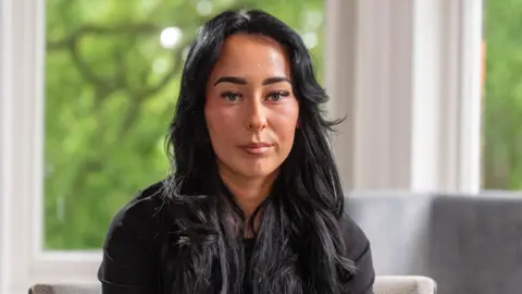 Kiera, dressed in a black hooded top, can be seen looking at the camera. She is seated and a garden can be seen in the window behind her.