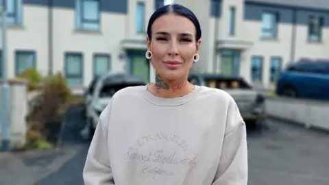 A woman, with black hair tied back, standing in front of a row of houses. She is wearing a cream coloured jumper and has white earrings. She has tattoos on her neck. 
