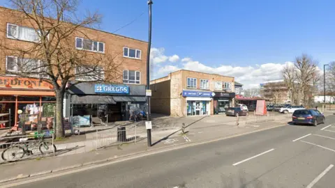 A Google maps image of a road and a row of shops, including a Greggs. There is a car driving on the road. 