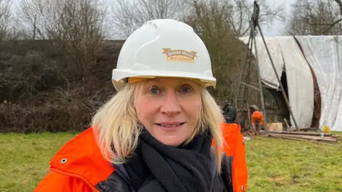 Lesley Carr wearing a bright orange high-vis jacket and white hard hat with the damaged bridge in the background largely covered in a white sheet.