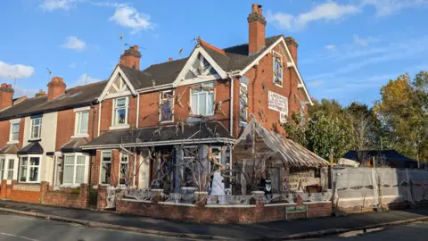 BBC A semi-detached house at the end of the street covered in Halloween decorations. Boards have been placed across windows and doors while skeletons can be seen climbing the walls.