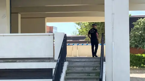 Henry Godfrey-Evans A uniformed police officer standing by a small outdoor staircase leading to the entrance of the tower block.