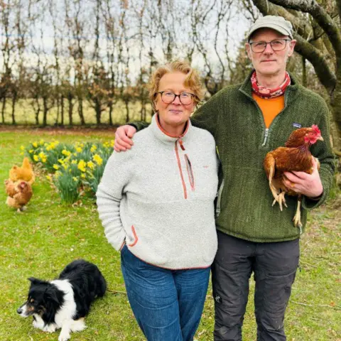 Marguerite Fleming and Frank Richards standing in a garden, he is holding a chicken, there are other chickens in the background, and a black and white collie dog, and daffodils and trees.