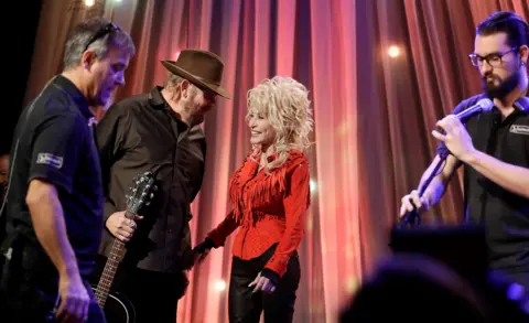Mark Humphrey/AP Dolly Parton talks and laughs with Hank Williams Jr on stage, while two stage crews work around them. She wears a bright red shirt with tassels. 