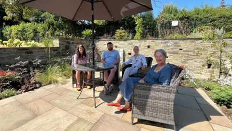 Ellen Knight/BBC Photograph of four people sat on chairs underneath a large grey parasol on a beige stone patio. Along the edges of the patio are borders with a variety of plants and flowers in them. The sky is blue and there is strong sunshine. 