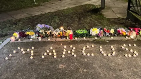 Small candles arranged to spell out the word Marcus on the ground. Floral tributes are behind them on the ground.