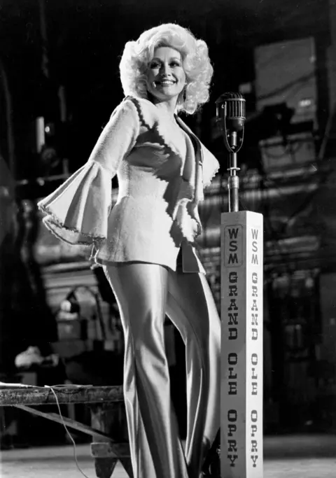 Nancy Barr Brandon/MediaPunch/Shutterstock Dolly Parton photographed at the Grand Ole Opry. She wears a suit with bell sleeves and smiles as she stands by a microphone 