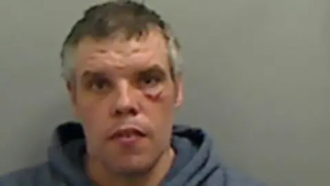 Paul Devaney has short grey hair and wears a blue sweatshirt. There is a bright red mark under his right eye. 