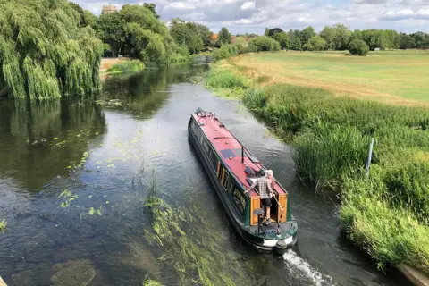 Sunshine Dan / BBC Weather Watchers A slightly raised view of a narrowboat in Bidford-on-Avon. The water seems fairly shallow, with plants and rocks visible beneath the surface. A field of grass is on drops to the right hand bank, while trees, including willows edge the left hand side