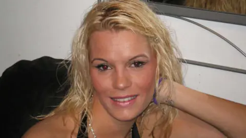 Head and shoulders shot of Kirsty. She is made up and has bleached blonde hair to her shoulders. She is wearing a black halter neck top and has a silver and sparkling stone necklace on. She is smiling slightly at the camera. There is the edge of a mirror behind her on a wall.