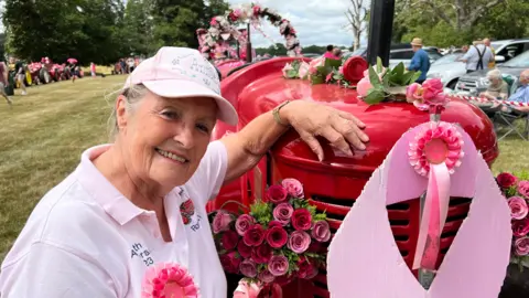 Mousumi Bakshi/BBC A woman stands next to a red tractor and smiles at the camera. She wears a pink cap and pink polo shirt. Her tractor is decorated with pink rosettes, red and pink roses and other pink floral arrangements. 