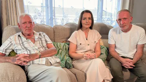 Three people on a beige sofa in a living room, with a window behind them