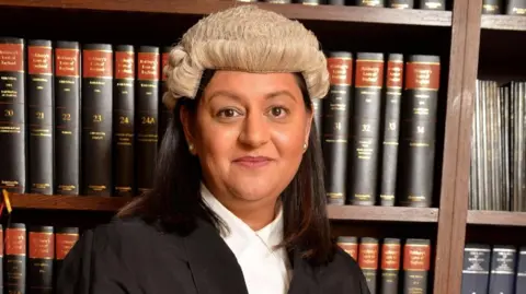 Sheriff Principal Aisha Anwar - she is in front of a bookcase filled with legal books, and has her sheriff's wig on. She has should length dark hair and is smiling at the camera.