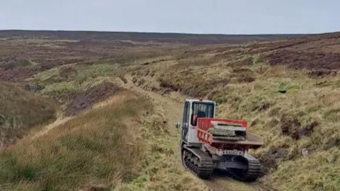 A grassy section of the Pennine Way in Bradford with a digger carrying flatstones 