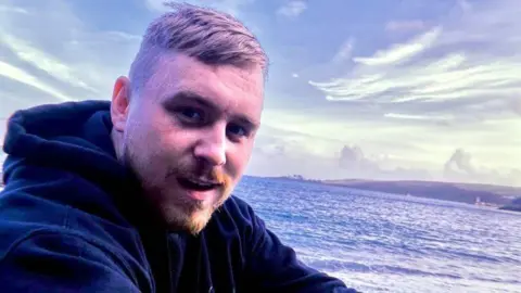 Family handout photo of Karol Zalewski sat along the coast with the sea behind him. He has a ginger goatee beard and is wearing a navy blue hoodie.