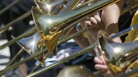 Getty Images Close-up of three gold trumpets being held by three people standing a line. Sunlight in shining on the instruments. 