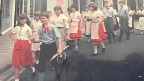 Children walking in two tows through a street, the girls are wearing white shirts and red skirts and the boys are wearing black trousers and light blue shorts wit red neckerchiefs. 