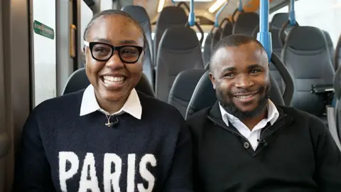 Uloma Igwe and Gabriel Enyi sitting on seats in a bus and laughing. Her black hair is pulled back from her face and she is wearing black framed glasses and a black jumper with Paris in white letters on its front and a white shirt collar sticking out of its crew neck. He has very short black hair and a black beard and is wearing a half-zip black jumper over a white shirt. 