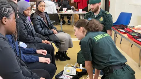 Young women sit watching as two paramedics demonstrate CPR on a dummy. The women are sat in a row in a community hall. One wears a brown hijab, with a black puffer coat, wool long dress and black lace up heeled boots, next to her a young women wears a grey hijab with a black collared puffer coat. There are two other women who's faces are just out of vision as they look away from the camera to watch the paramedics. The paramedics both wear forest green uniform, which has the word 'Ambulance' across the back. A female paramedic with brown hair tied back in a ponytail has both her palms pressing down on a dummy's chest demonstrating CPR, while her male colleague can be seen in the background looking at the young women.