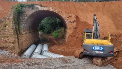 Severn Valley Railway A red earth tunnel with three metal tubes running through it and a yellow and black mechanical digger in the foreground
