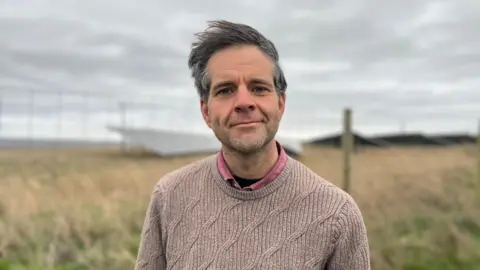 David Woricker looking into camera with the solar farm behind him. He has dark hair which is being blown in the wind, is wearing a light brown jumper and pink shirt.