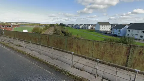 Google A view over a bridge with houses and a hill in the background. The sky is blue with a few clouds.