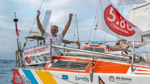 Arms aloft in triumph, grey-haired Adam Waugh arrives in Antigua aboard his white, red, blue and yellow 19ft (5.8m) yacht called Little Wren, adorned in flags and sponsorship logos, travelling 28,000 nautical miles in the inaugural Mini Globe Race.