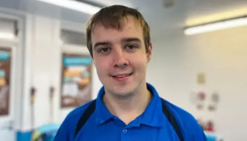 A man with short blonde hair is smiling towards the camera. He is wearing a blue polo shirt. 