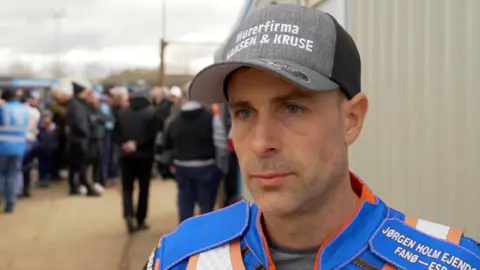 Niels-Kristian Iversen is in his blue racing jacket and wearing a black and grey baseball cap. He is standing outside and there are people huddled behind him.