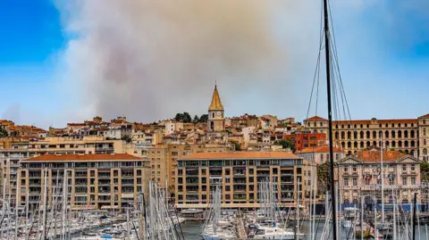 A fire currently burning in the hills north of Marseille, in the area of Les Pennes-Mirabeau, is producing thick smoke visible from the Vieux-Port. A large grey smoke cloud can be seen above orange-roofed buildings by the waterside, with small boats in the foreground.