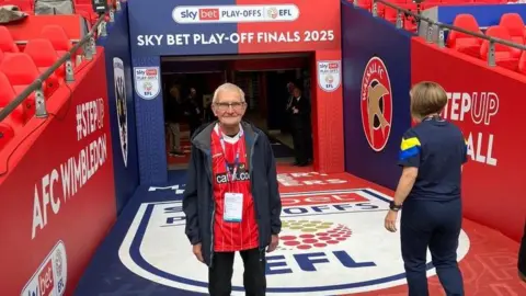 Gary Edwards standing in the pitchside tunnel on his VIP tour of Wembley. He is wearing a red football top and blue jacket and has a lanyard around his neck. He has grey hair and is wearing glasses. A woman is walking the other way beside him.