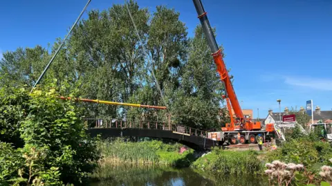 BBC Arch-shaped bridge over a river, with a crane positioning itself, ready for removal.