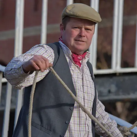Kev Maslin A man with a green flat cap on, grey waistcoat, white and red checked shirt and a rope in his hands