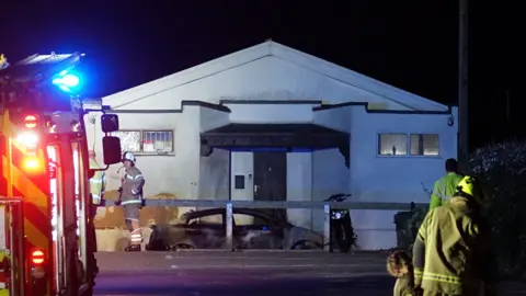 Eddie Mitchell A white building is surrounded by firefighters at night.