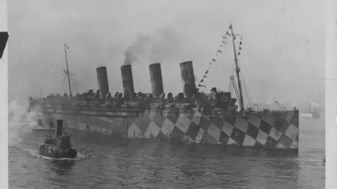 Getty Images A black and white picture of a World War One ship in water. It has four large funnels that are blowing smoke. A mast as bunting on it. The ship is painted in a black and white checkerboard pattern