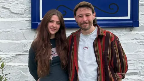 A young woman with long brown hair in a dark blue top and a many with brown hair and a beard in a striped shirt with a white shirt underneath