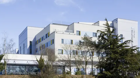 The Beach building at Royal Bournemouth Hospital, as seen from distance. The white, multi-storey, compartmented building sits on a grassy bank behind trees.
