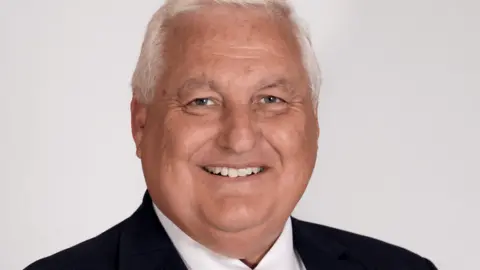 Barry Faudemer is smiling and is wearing a suit. There is a white background behind him.