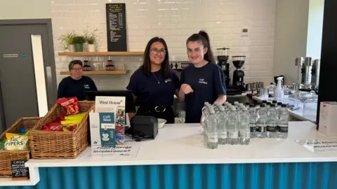 Ed Hanson/BBC Three women standing behind the counter at the cafe which is blue at the front with a white counter top. Two of the women are standing centrally and both have brown hair and are smiling. There are bottles of water in front of them to the right as well as brochures and baskets full of crisps to the left. There is a sink and coffee machine behind them. There is another woman standing further back to the left and is wearing glasses.