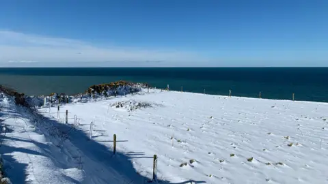 Snow on a field on the edge of a sand cliff with the Irish Sea below.