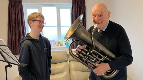 Neythen is standing in a living room next to Brian Routledge, who is holding a euphonium. They are both smiling.