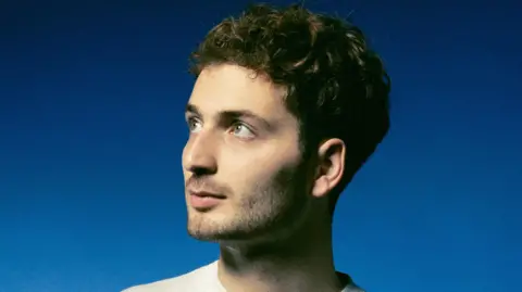 Charlie Floyd Charlie Floyd is looking towards the sky with a neutral expression with a navy blue background behind him. He is wearing a white t-shirt and has brown curly hair with light stubble.