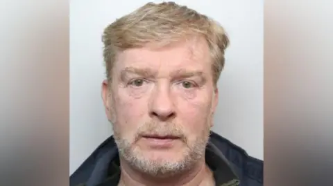 gmp Mugshot of Aron Traynor who has short sandy-coloured hair and beard. He wears a dark jacket