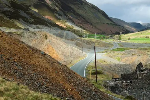 Jon Pountney Pile of different coloured rubble and stone at the side of the road leading through a valley